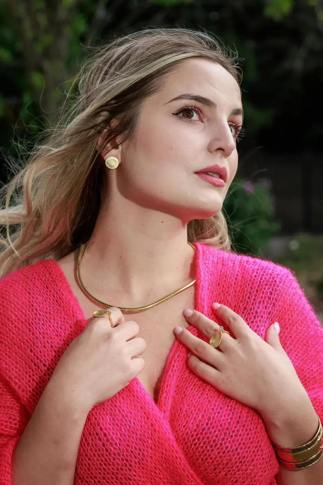 Woman wearing gold jewelry set with earrings, necklace, rings, and bracelet, in pink sweater
