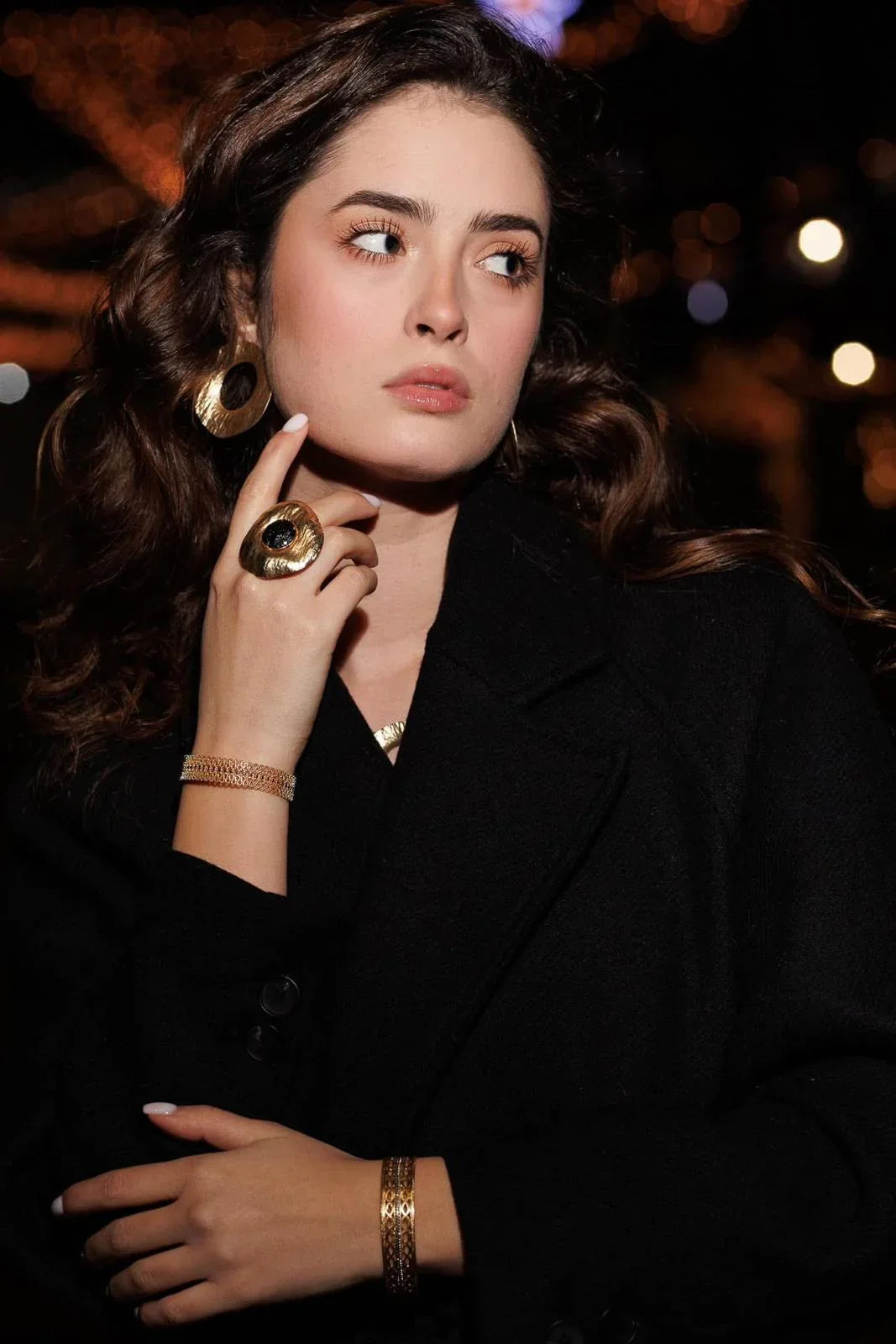 Woman in black coat wearing statement gold jewelry, including ring, earrings, and bracelets