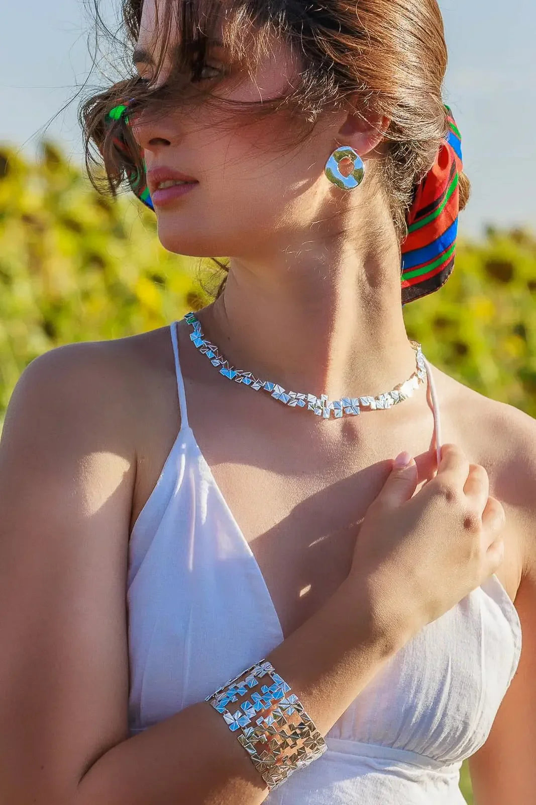 Woman outdoors wearing silver geometric necklace, cuff bracelet, round earrings, and white dress