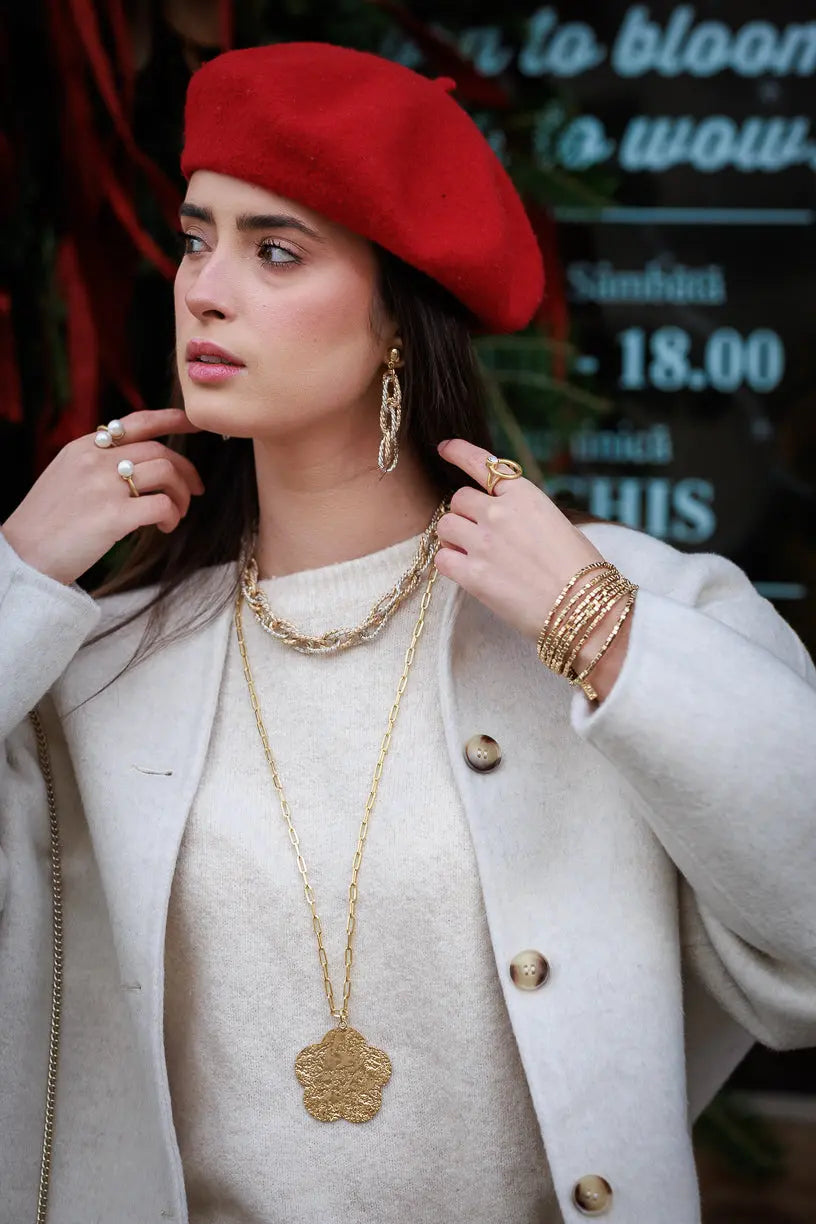 Woman wearing a red beret, cream coat, and layered gold jewelry including necklace, earrings, rings, and bracelets